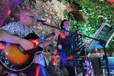 Miniature DJ Pézilla-la-Rivière (Pyrénées-Orientales) - Cleanjack #2 Un duo musical se produit sur une scène intérieure avec guitare et chant, devant un public assis.