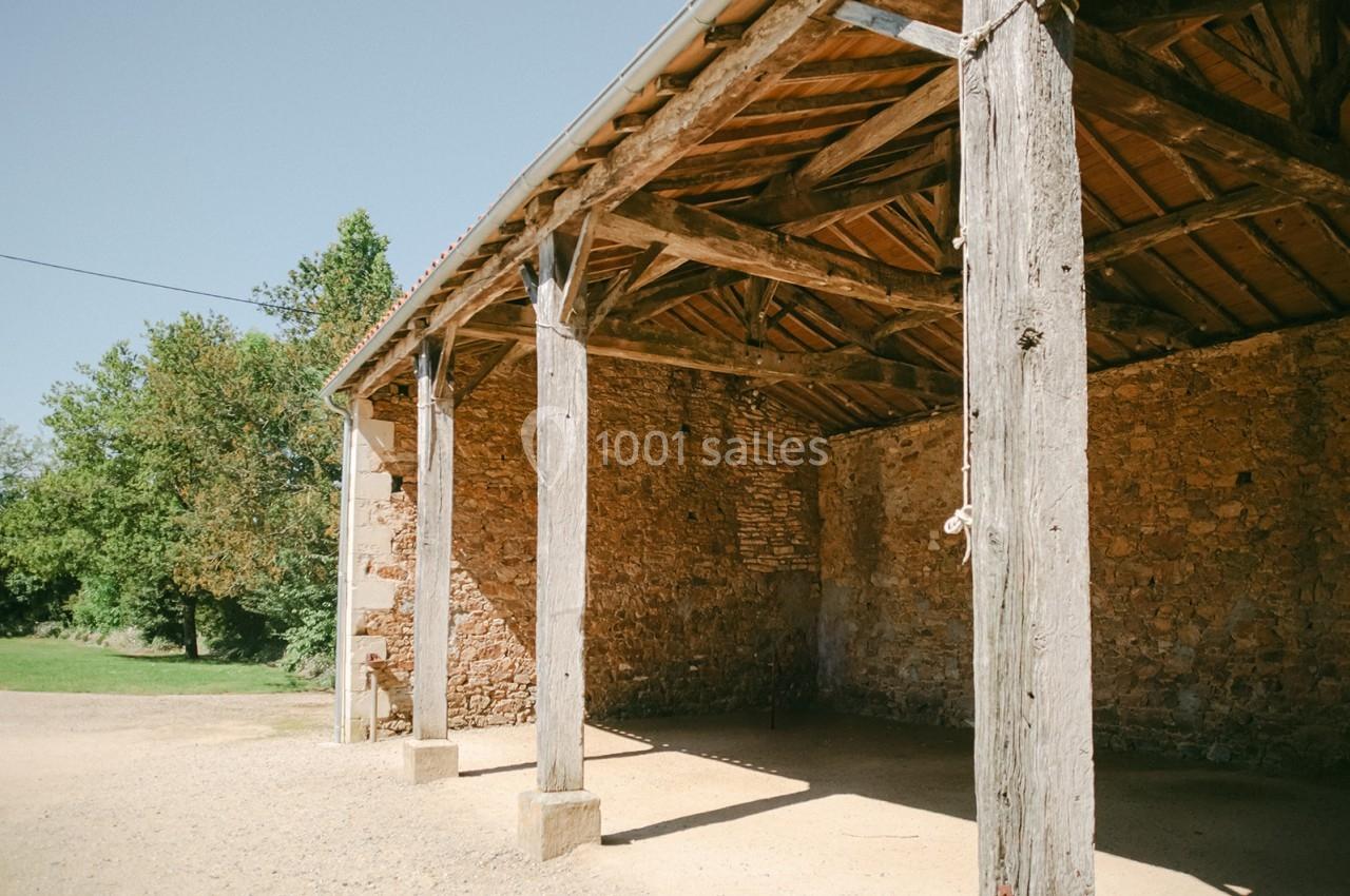 Abri en bois avec toit en tuiles, soutenu par des poteaux, situé près d'un mur en pierre dans un environnement rural.