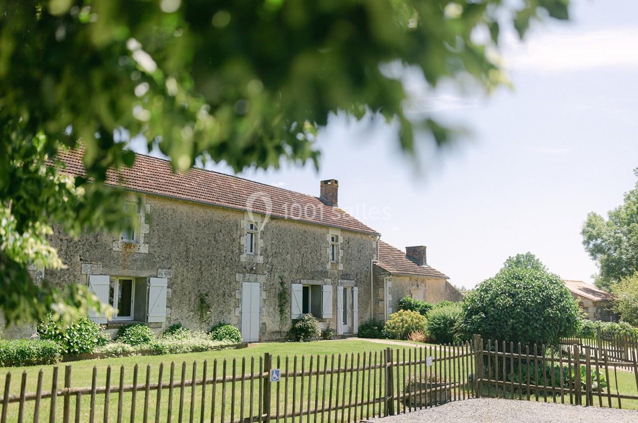 Maison en pierre avec volets blancs entourée de verdure, vue à travers des branches et une clôture en bois.