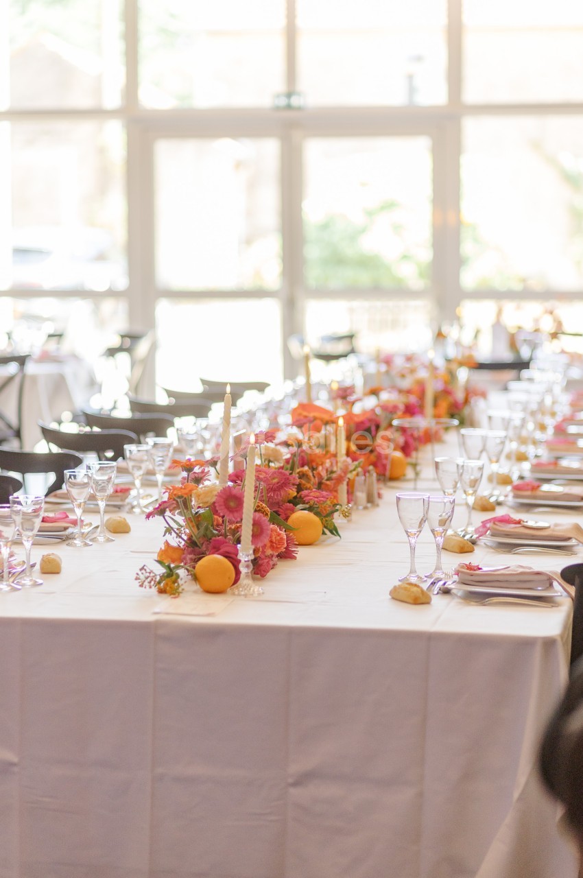 Table dressée avec une nappe blanche, décorée de fleurs colorées, bougies et fruits, dans une salle lumineuse.