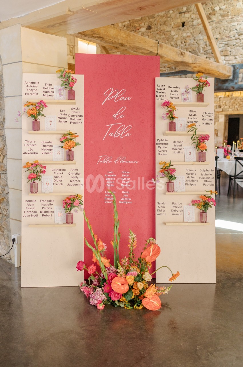 Panneau de plan de table de mariage avec fleurs colorées, disposé dans une salle en pierre avec poutres apparentes.