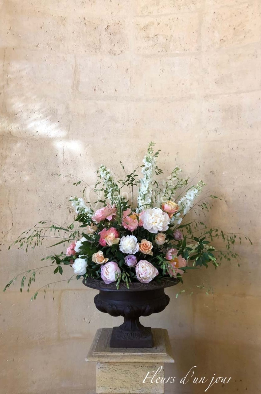 Arrangement floral avec des roses, pivoines et delphiniums dans un vase en métal sombre sur un socle en pierre beige.