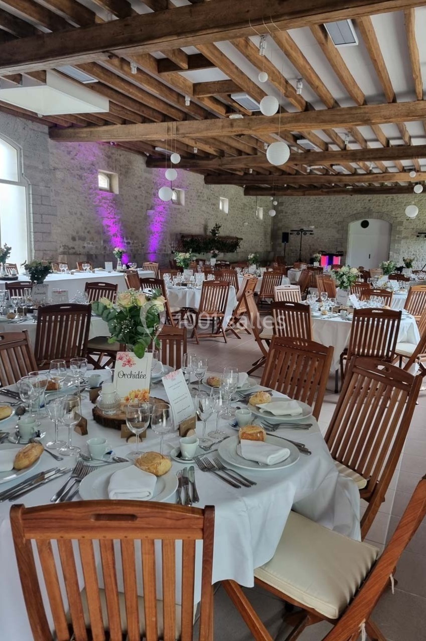 Salle de réception décorée avec des tables rondes dressées, chaises en bois et éclairage tamisé.