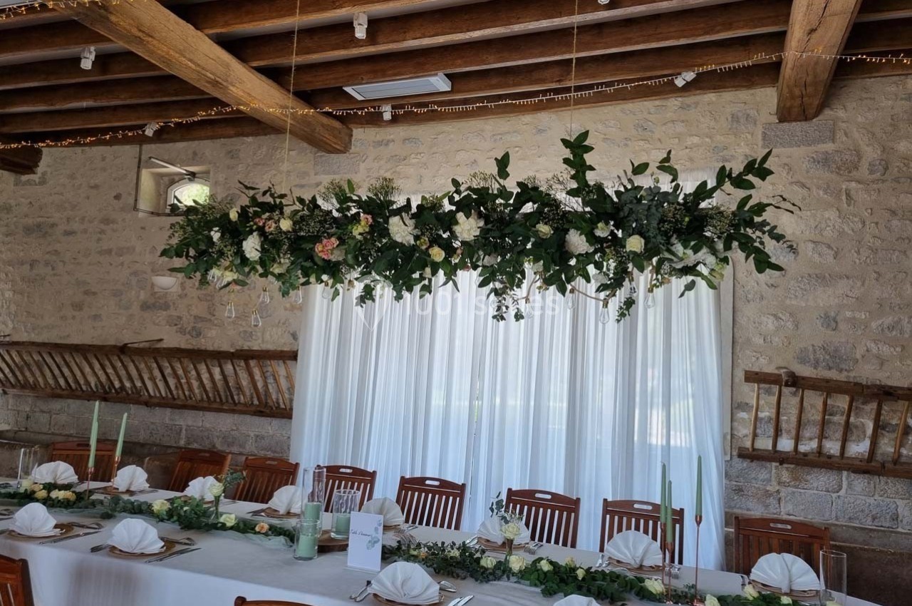 Décoration florale suspendue au-dessus d'une table de réception dressée dans une salle en pierre avec poutres apparentes.