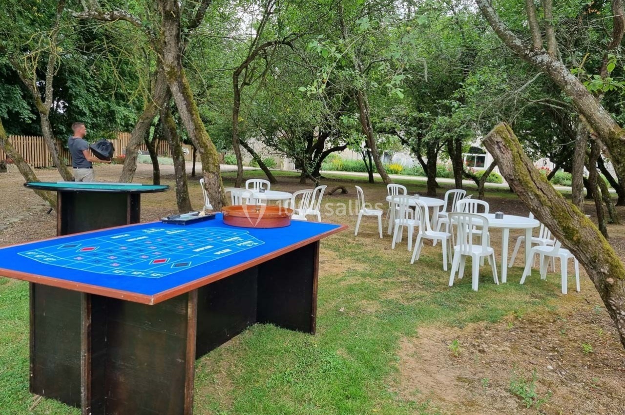 Tables de jeu et chaises blanches disposées dans un jardin arboré avec un homme en arrière-plan.