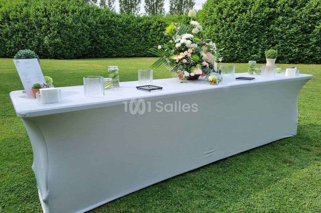 Table rectangulaire blanche dressée en extérieur, décorée de fleurs, bougies et petits objets sur une pelouse verdoyante.