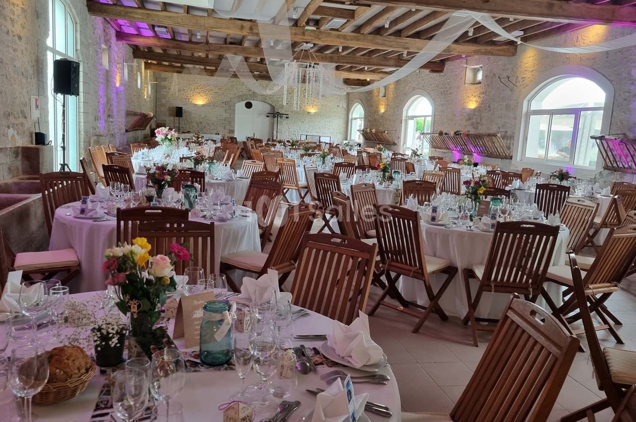 Salle de réception décorée avec des tables rondes dressées, chaises en bois et éclairage tamisé.