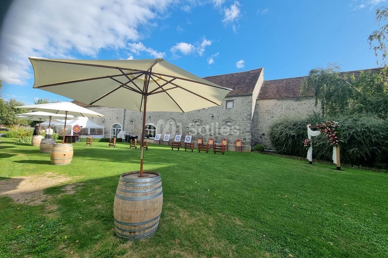 Cour extérieure avec pelouse, parasols sur tonneaux, chaises alignées et bâtiment en pierre sous un ciel dégagé.