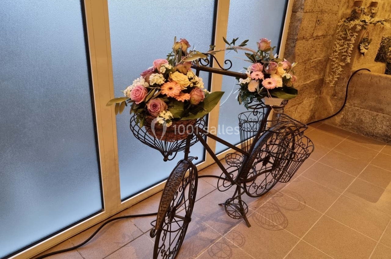Vélo décoratif en métal noir avec des paniers garnis de fleurs roses et blanches, placé devant une fenêtre givrée.