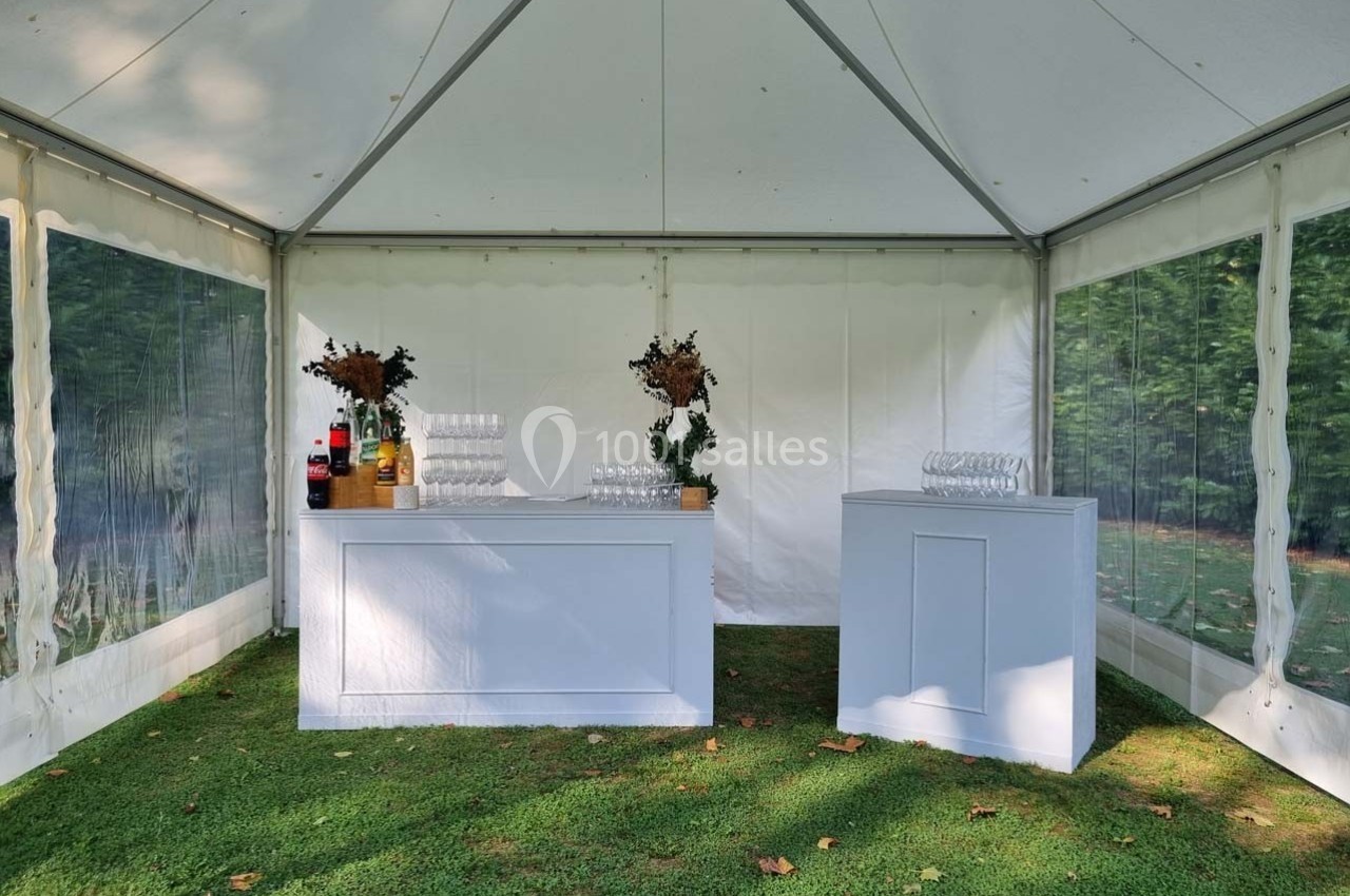 Bar blanc sous une tente, avec verres, bouteilles et décorations florales, installé sur une pelouse.