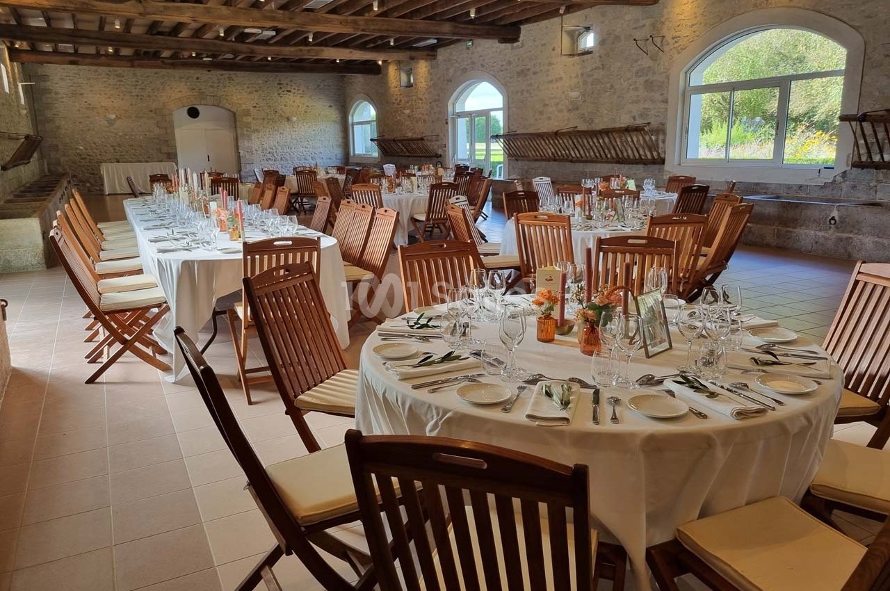Salle de réception lumineuse avec tables rondes et rectangulaires dressées, chaises en bois et murs en pierre apparente.