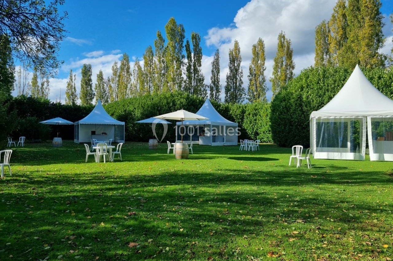 Espace extérieur avec pelouse, tentes blanches, tables et chaises sous un ciel partiellement nuageux.