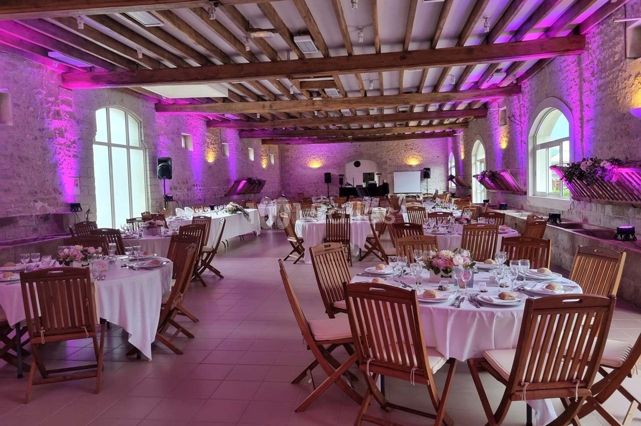 Salle de réception en pierre avec tables rondes dressées, chaises en bois et éclairage rose diffus.