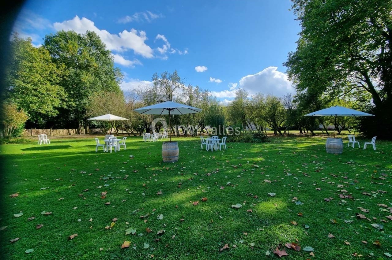 Chaises et tables blanches disposées sur une pelouse avec des parasols, entourées d'arbres sous un ciel dégagé.