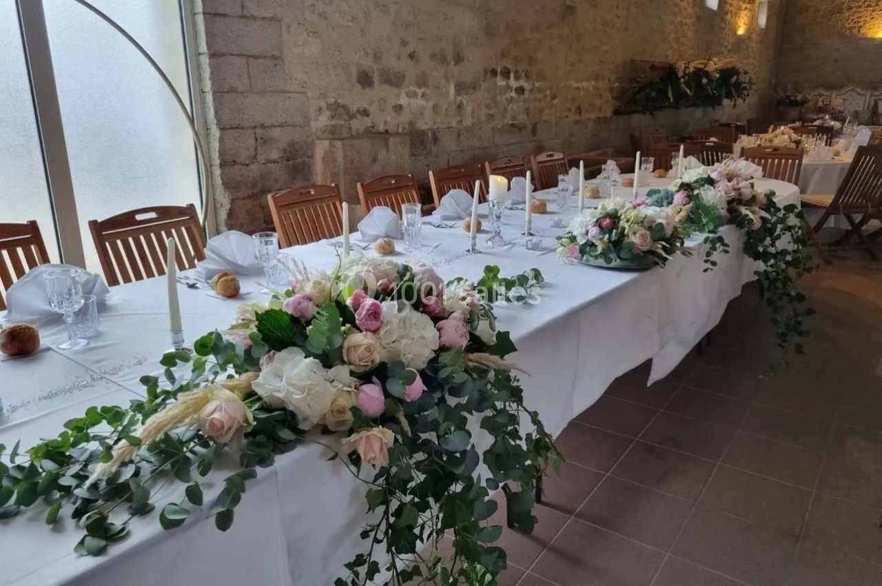 Table de réception décorée avec des fleurs blanches et roses, nappes blanches et chaises en bois dans une salle en pierre.