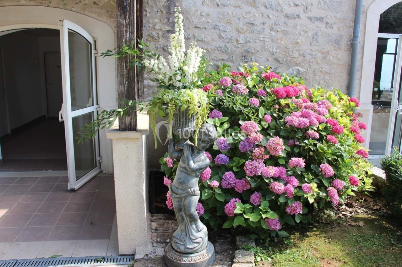 Statue décorative portant un vase fleuri devant un mur en pierre, près d'hortensias roses et d'une porte ouverte.