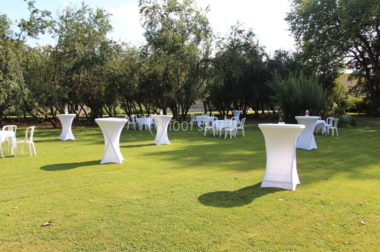 Tables hautes et chaises blanches disposées sur une pelouse verdoyante, entourées d'arbres.
