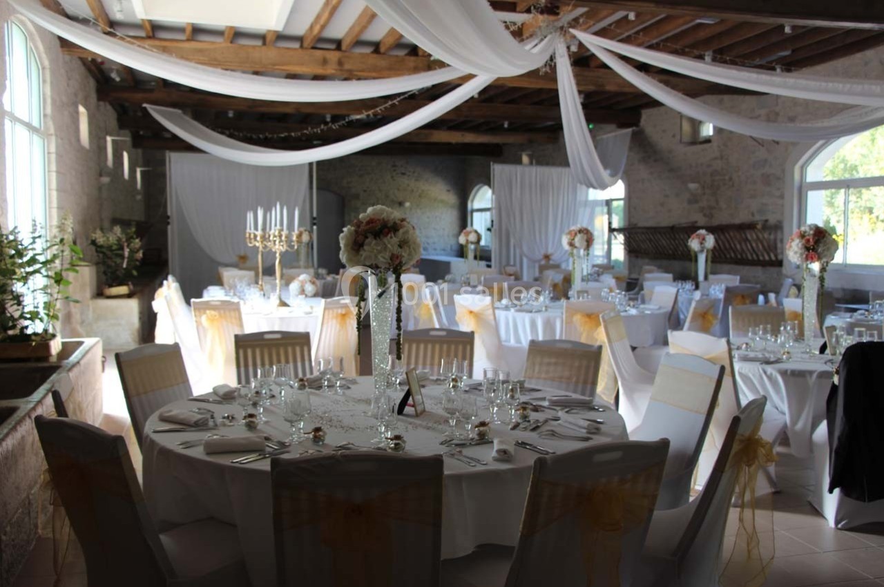 Salle de réception décorée avec des tables rondes, nappes blanches, chaises ornées de nœuds dorés et centres de table…