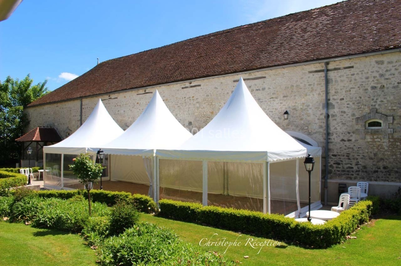 Tentes blanches installées devant un bâtiment en pierre, entourées de haies et d'un jardin bien entretenu.