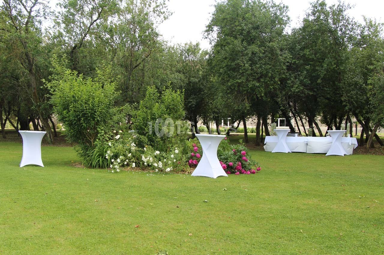 Jardin avec pelouse, tables hautes blanches et parterre de fleurs colorées entouré d'arbres.