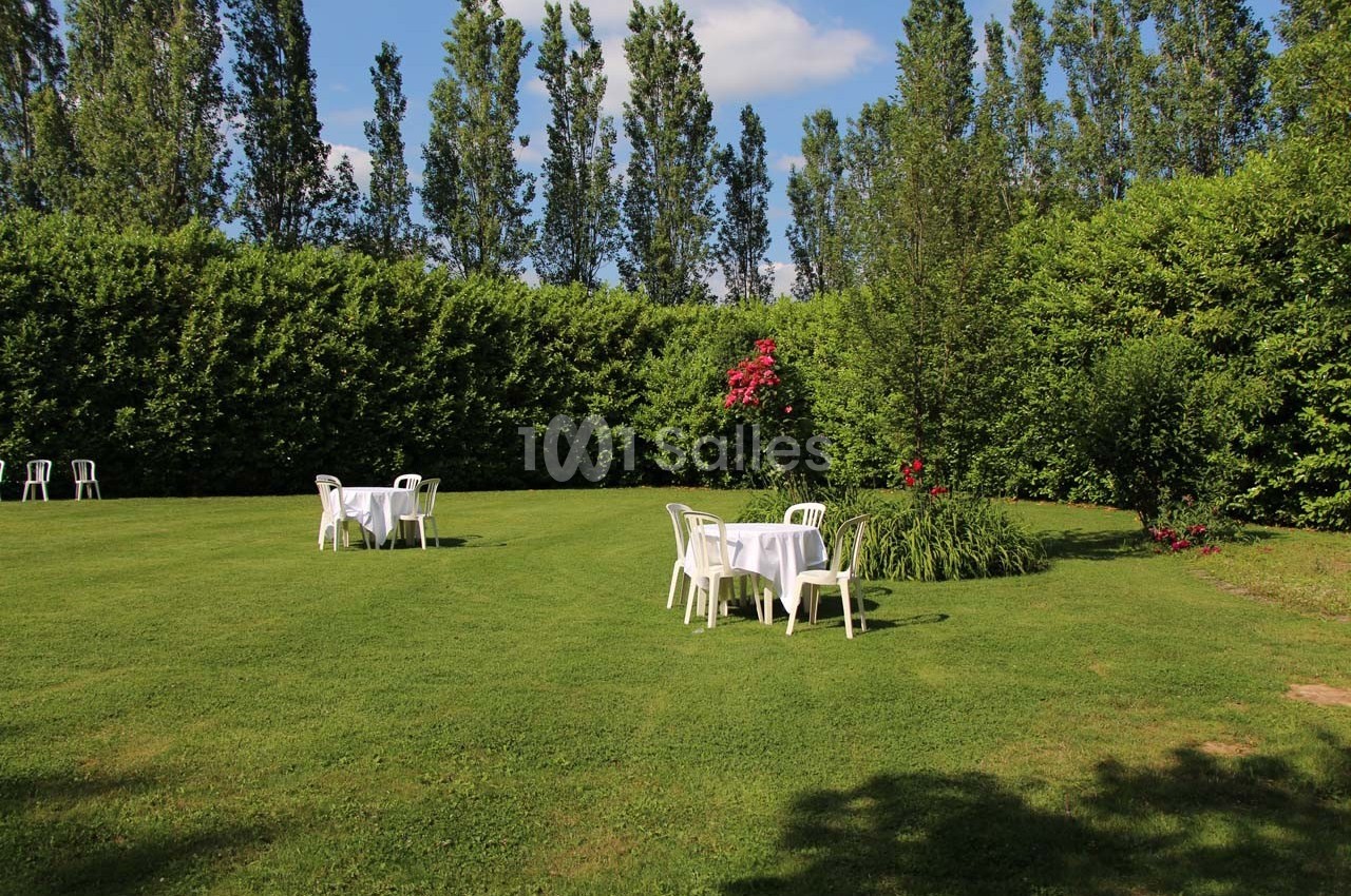 Tables blanches et chaises en plastique disposées sur une pelouse entourée de haies et d'arbres sous un ciel dégagé.