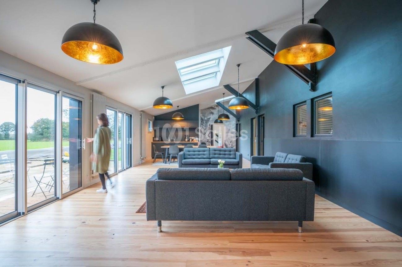 Salon moderne avec canapés gris, parquet clair, grandes baies vitrées et cuisine ouverte en arrière-plan.