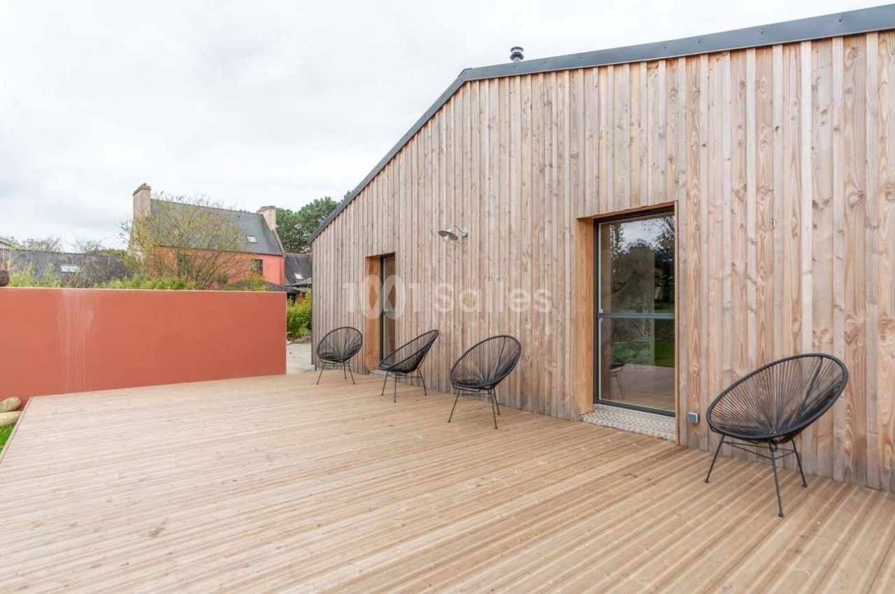 Terrasse en bois avec des chaises noires, adossée à une maison en bardage bois, sous un ciel nuageux.
