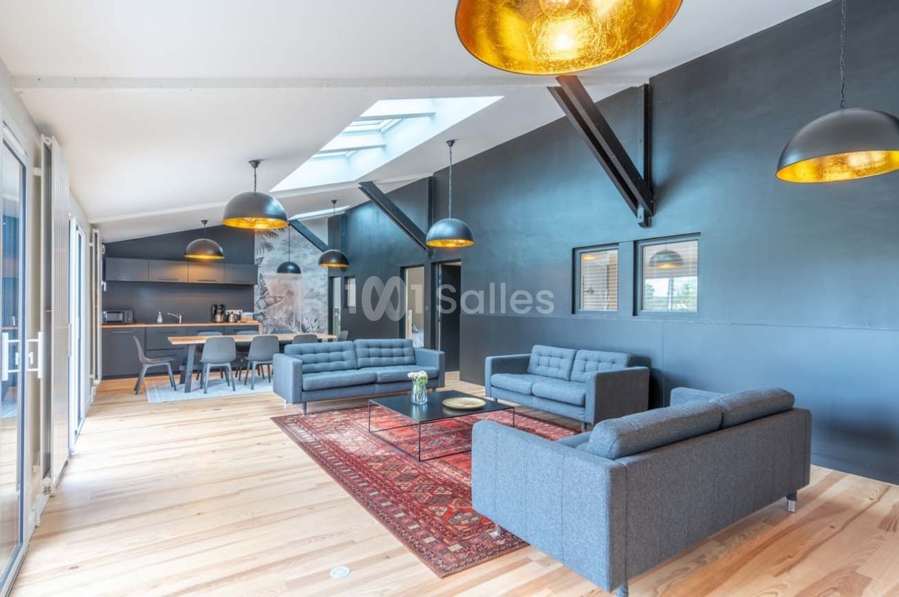 Espace de vie lumineux avec canapés gris, tapis rouge, cuisine ouverte et grandes suspensions dorées.