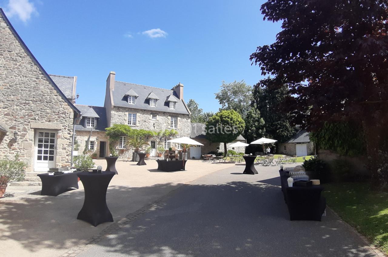 Cour d’un bâtiment en pierre avec tables hautes noires, parasols blancs et végétation sous un ciel ensoleillé.