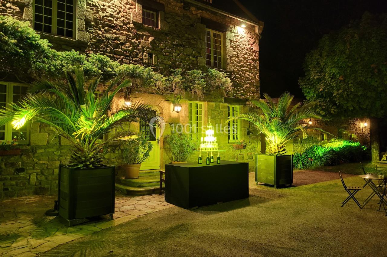 Façade en pierre éclairée de nuit, avec palmiers illuminés et table décorée de bouteilles sur une terrasse.