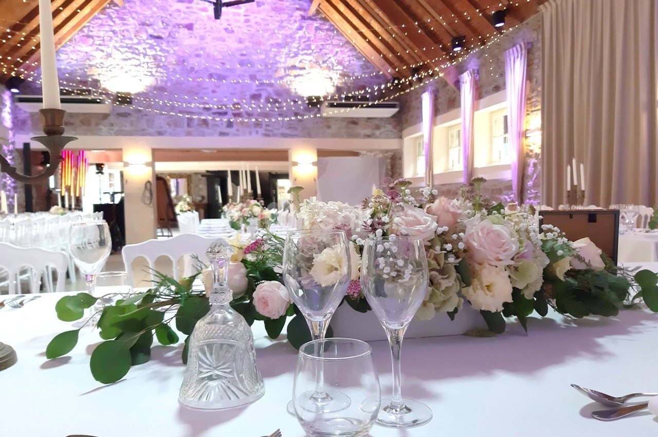 Salle de réception décorée avec des guirlandes lumineuses, des fleurs pastel et une table dressée élégamment.