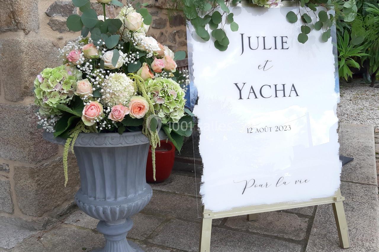 Panneau de mariage décoré de fleurs blanches et roses, placé à côté d'une jardinière fleurie devant un bâtiment en pierre.