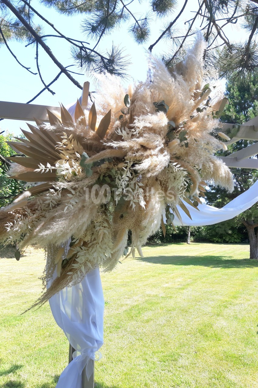 Arrangement floral avec des herbes de la pampa et feuillages, fixé à une structure en bois dans un jardin ensoleillé.