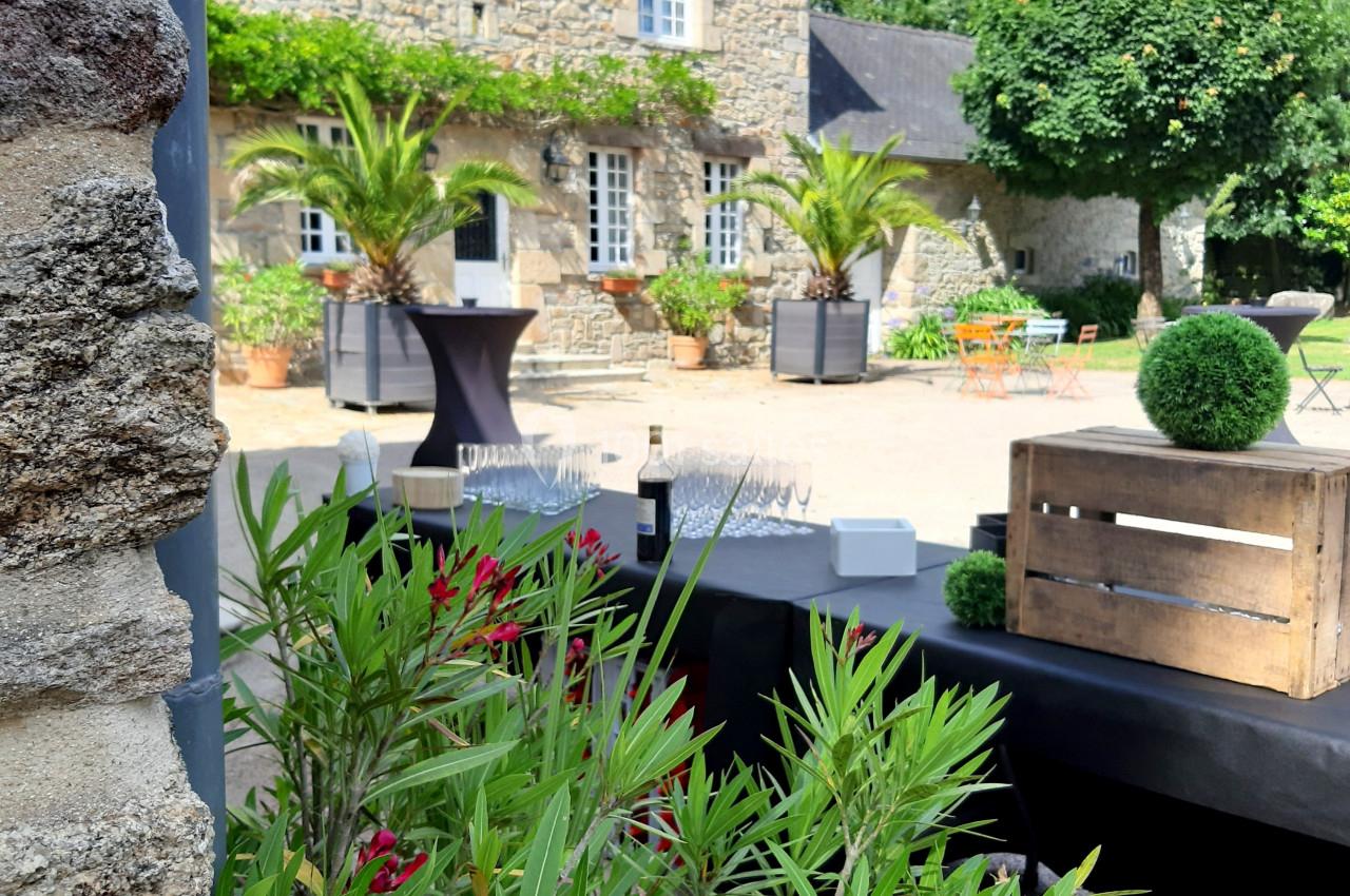Cour d'une maison en pierre avec des plantes, des tables hautes et des caisses en bois, sous un ciel partiellement nuageux.