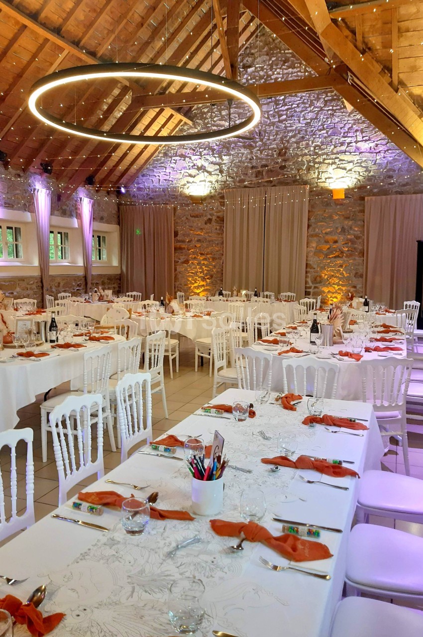 Salle de réception avec tables dressées, nappes blanches, serviettes orange, chaises blanches et éclairage chaleureux.