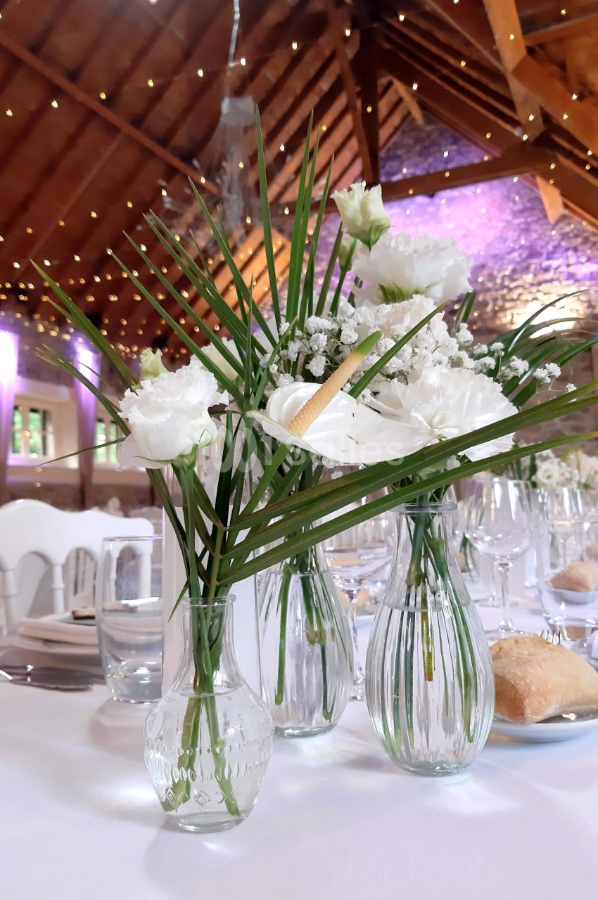 Vases en verre contenant des fleurs blanches et feuillages, disposés sur une table dressée dans une salle avec poutres…