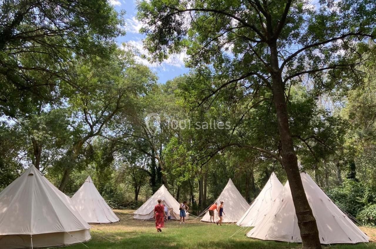 Tentes blanches installées dans une clairière ombragée, avec des personnes se déplaçant entre les arbres.