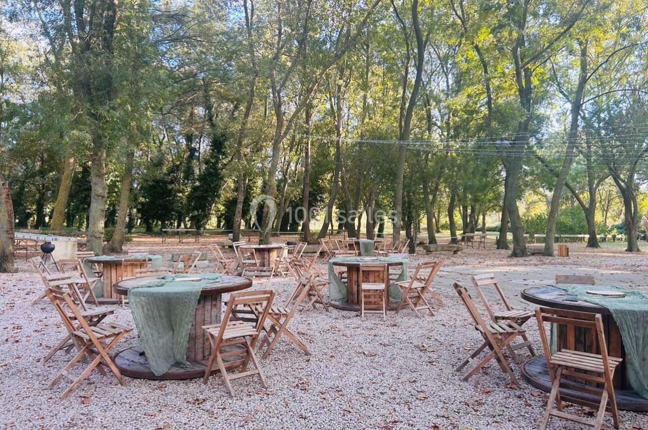 Tables et chaises en bois disposées en extérieur sur un sol gravillonné, entourées d'arbres dans un cadre naturel.