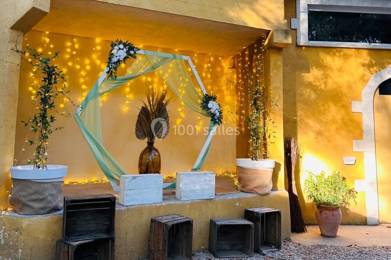 Décoration extérieure avec guirlandes lumineuses, voilage vert, plantes en pots et caisses en bois devant un mur jaune.