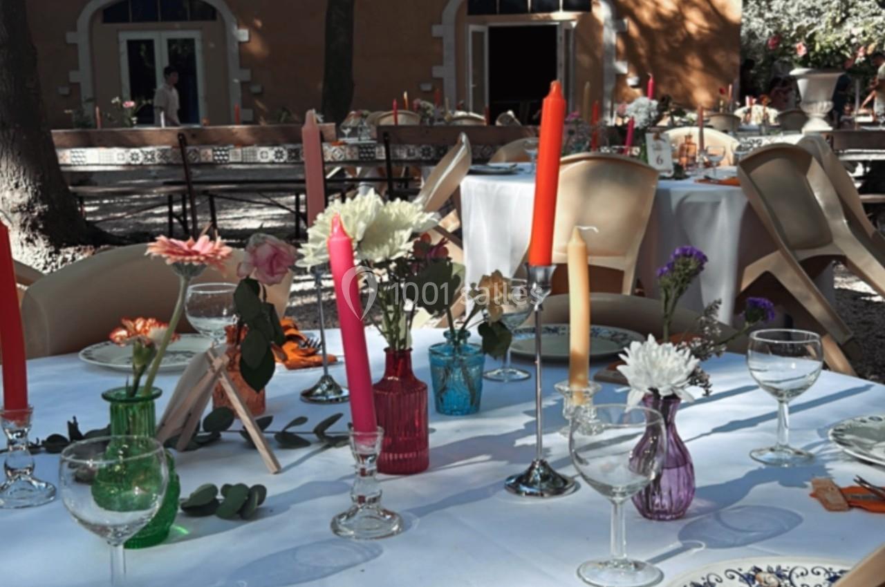 Table dressée en extérieur avec nappes blanches, bougies colorées, vaisselle et décorations florales sous des arbres.
