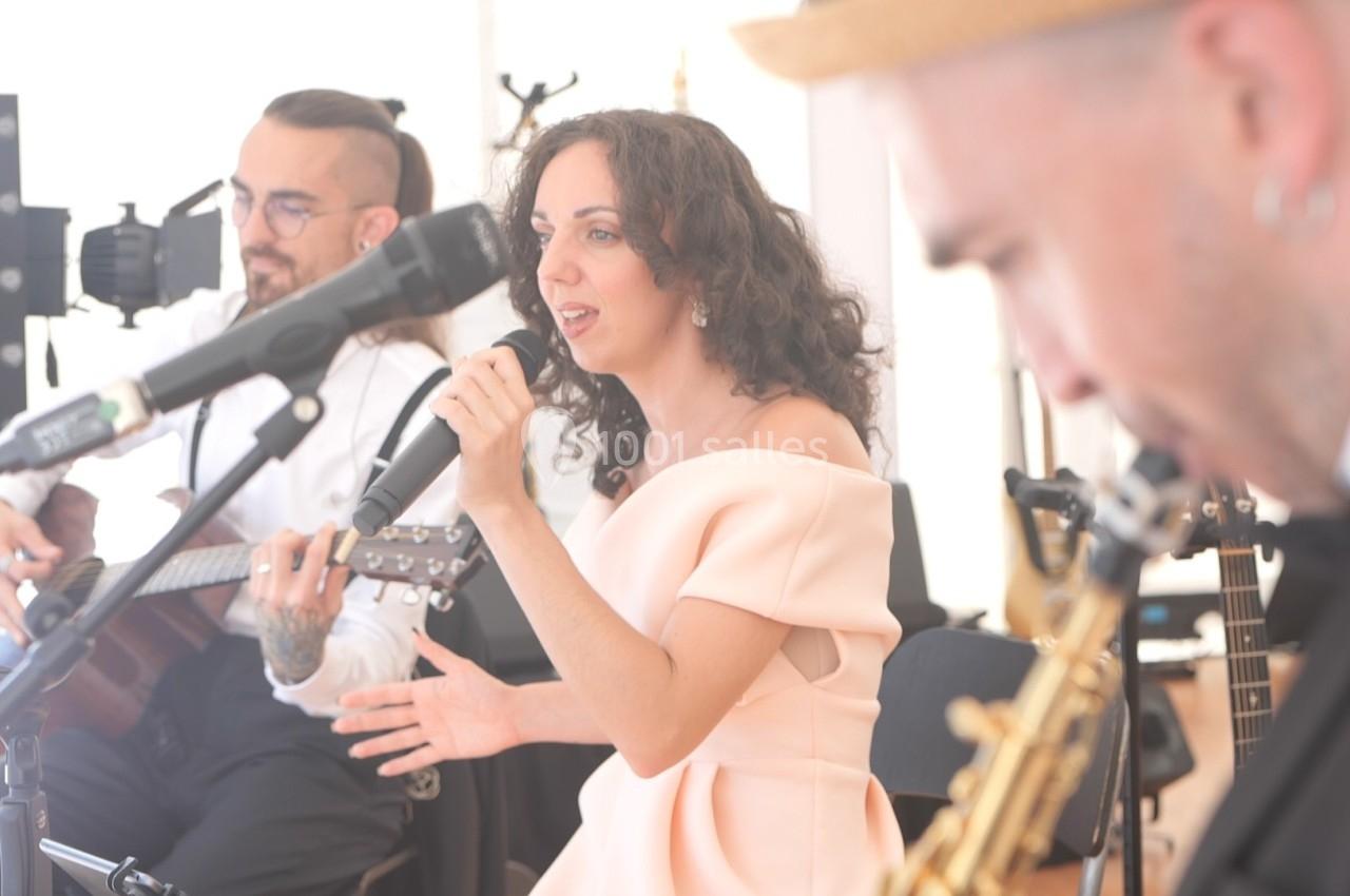 Une chanteuse interprète une chanson au micro, accompagnée de musiciens jouant de la guitare et du saxophone.