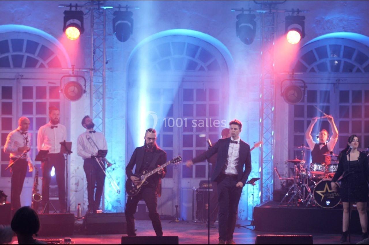 Groupe de musiciens et chanteurs sur scène, éclairés par des lumières colorées, jouant devant un public.