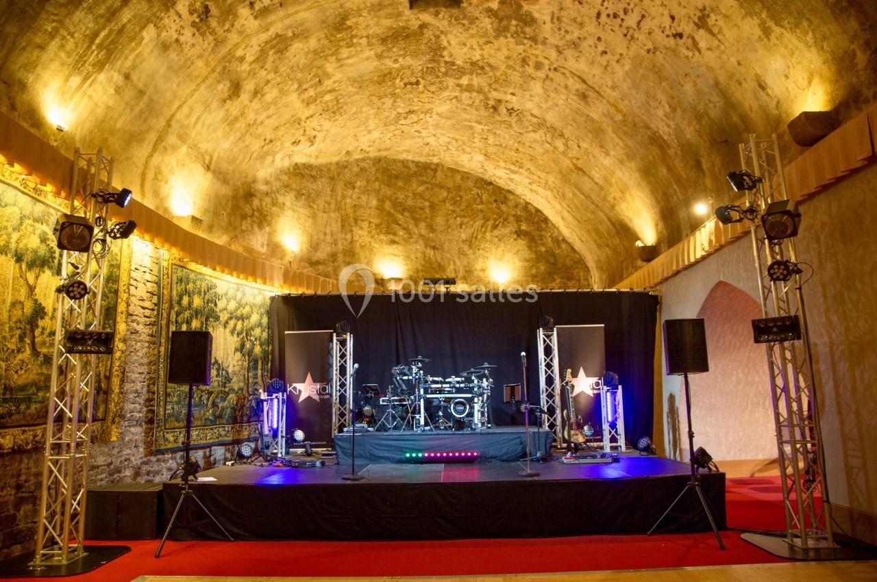 Scène de concert éclairée avec une batterie, installée dans une salle voûtée aux murs en pierre et tapisseries.