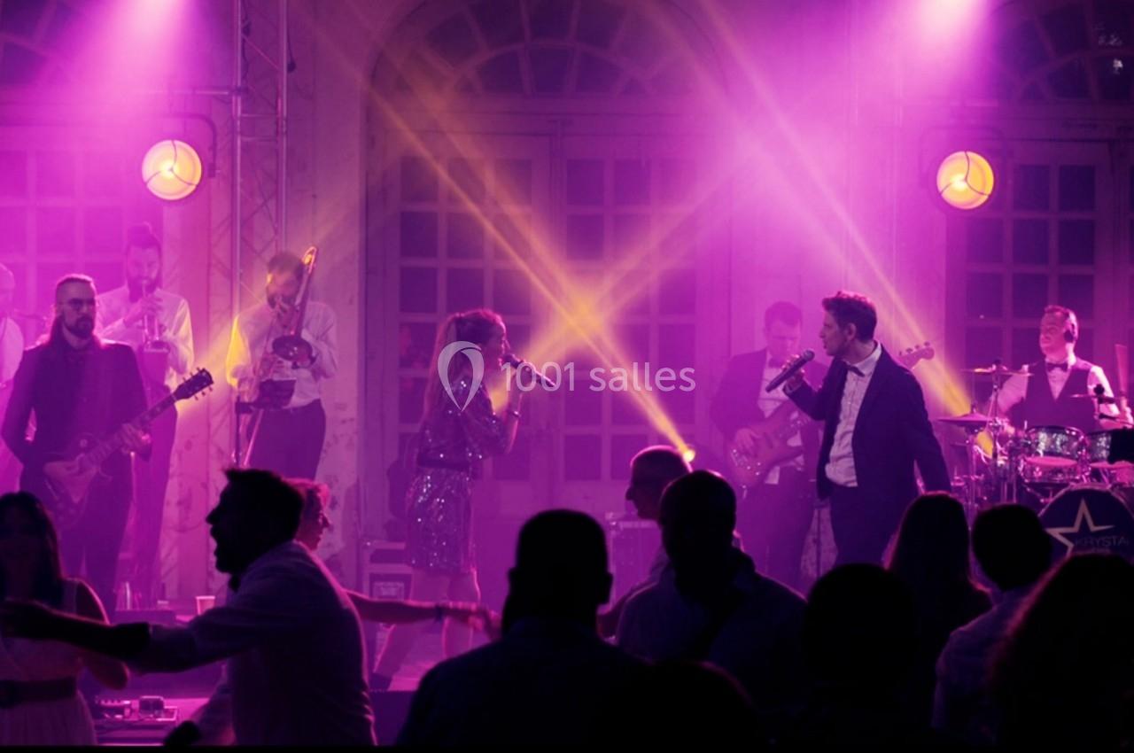 Groupe de musiciens et chanteurs se produisant sur scène sous des éclairages violets, avec un public dansant au premier plan.