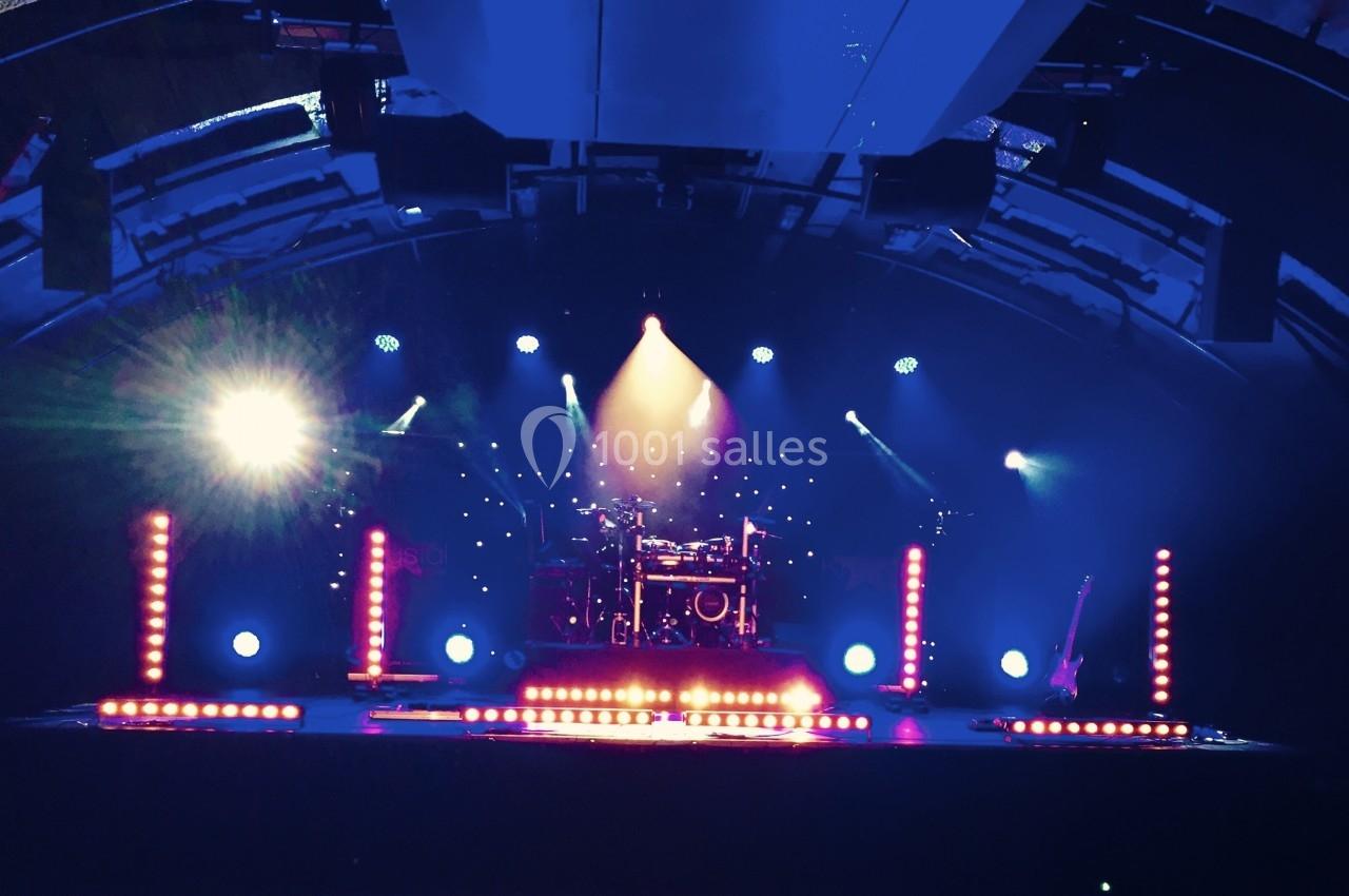 Scène de concert vide éclairée par des projecteurs colorés, avec une batterie et une guitare en arrière-plan.