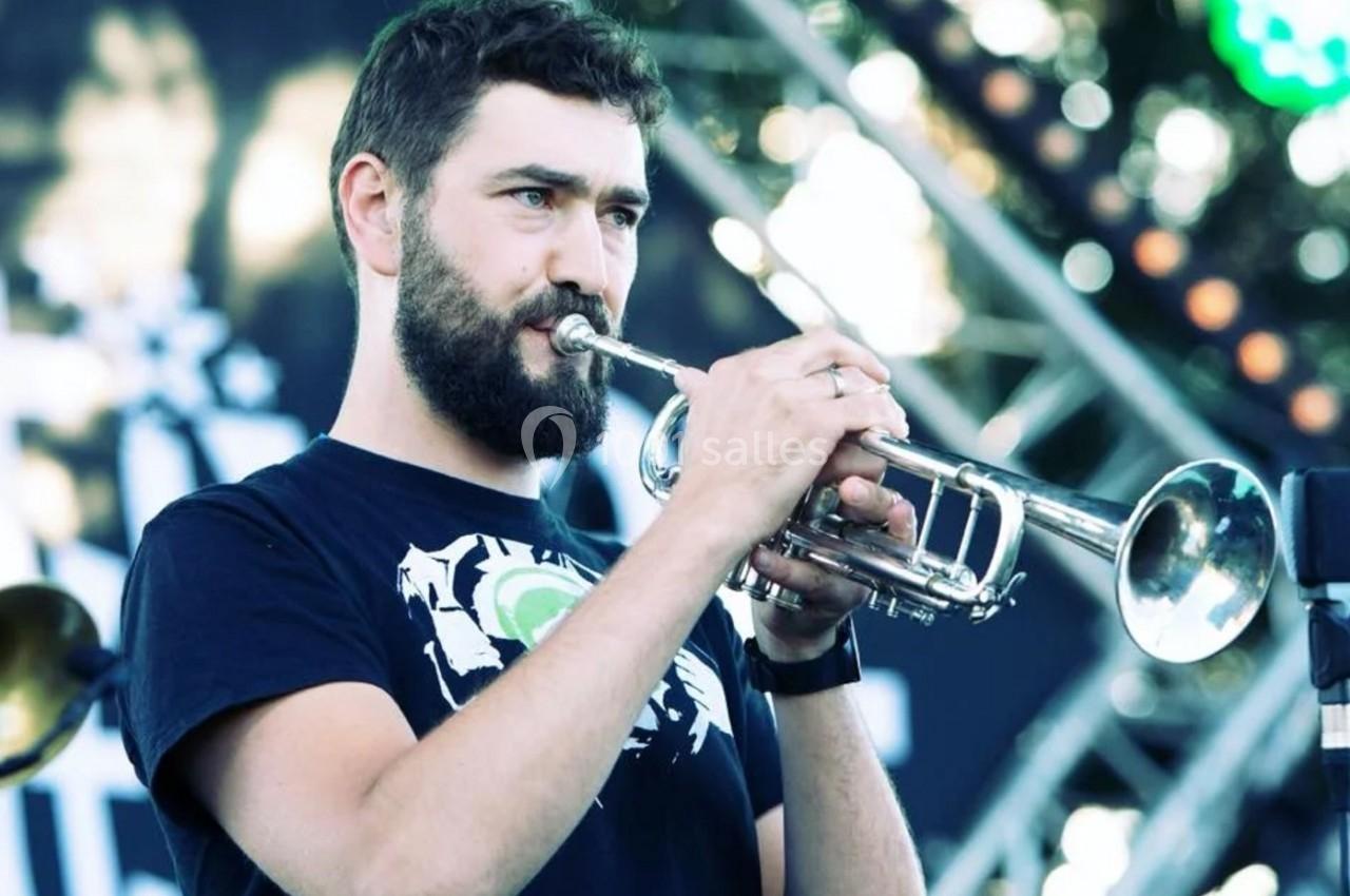 Un homme barbu joue de la trompette en extérieur, avec une scène et des lumières floues en arrière-plan.