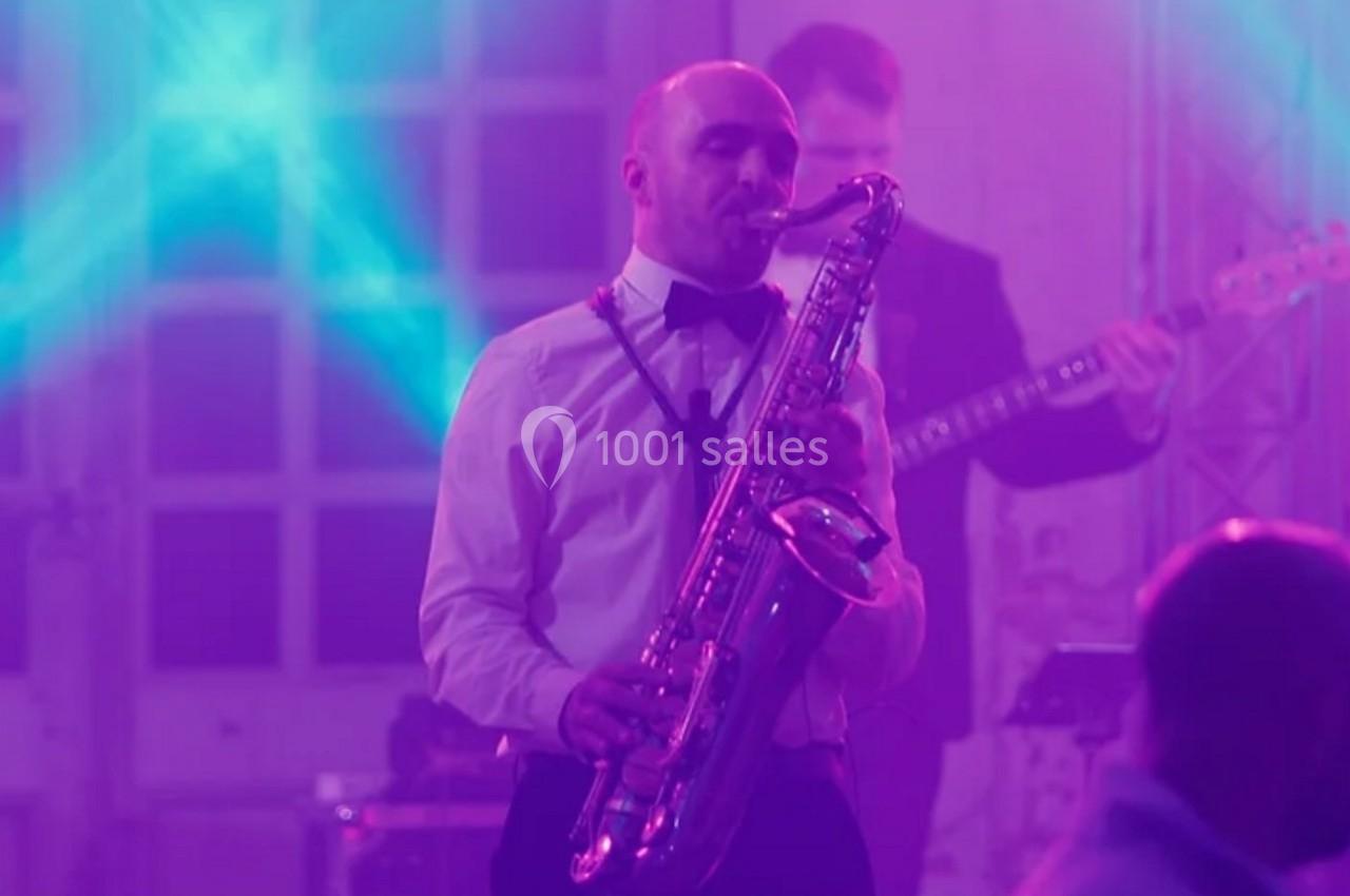 Un musicien joue du saxophone sur une scène éclairée de lumières violettes, accompagné d'autres artistes en arrière-plan.