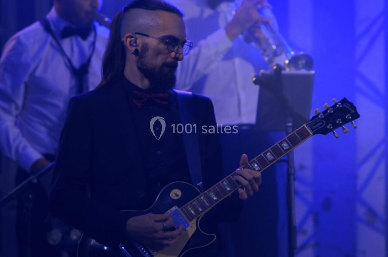 Un guitariste en costume joue sur scène sous un éclairage bleu, accompagné de musiciens en arrière-plan.