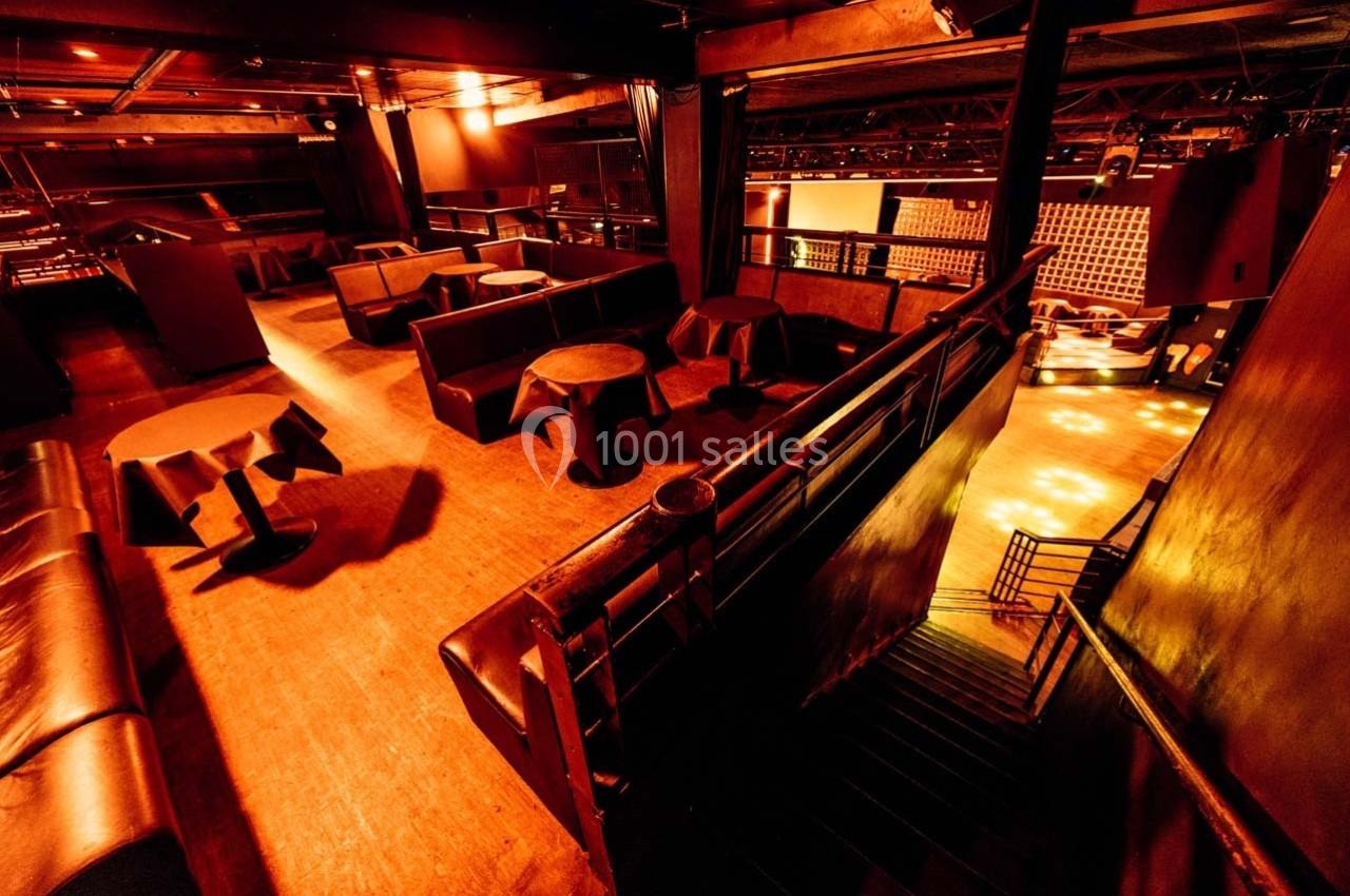 Salle de concert ou club avec éclairage tamisé, tables rondes, banquettes et escalier menant à une piste éclairée.