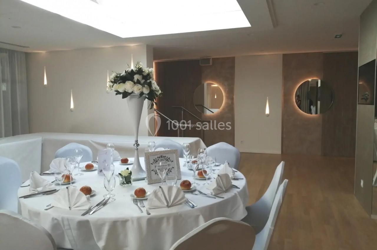 Salle de réception élégante avec table ronde dressée, nappes blanches, serviettes pliées et centre de table floral.